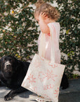 Child holding a tote bag with floral designs next to a black dog against a green leafy background