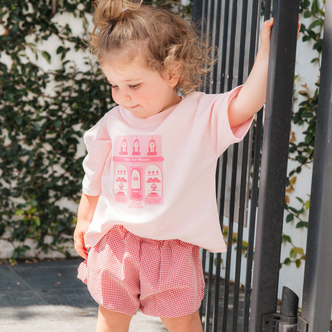 Child wearing a pink t-shirt with graphics and red checkered shorts standing next to a black gate.