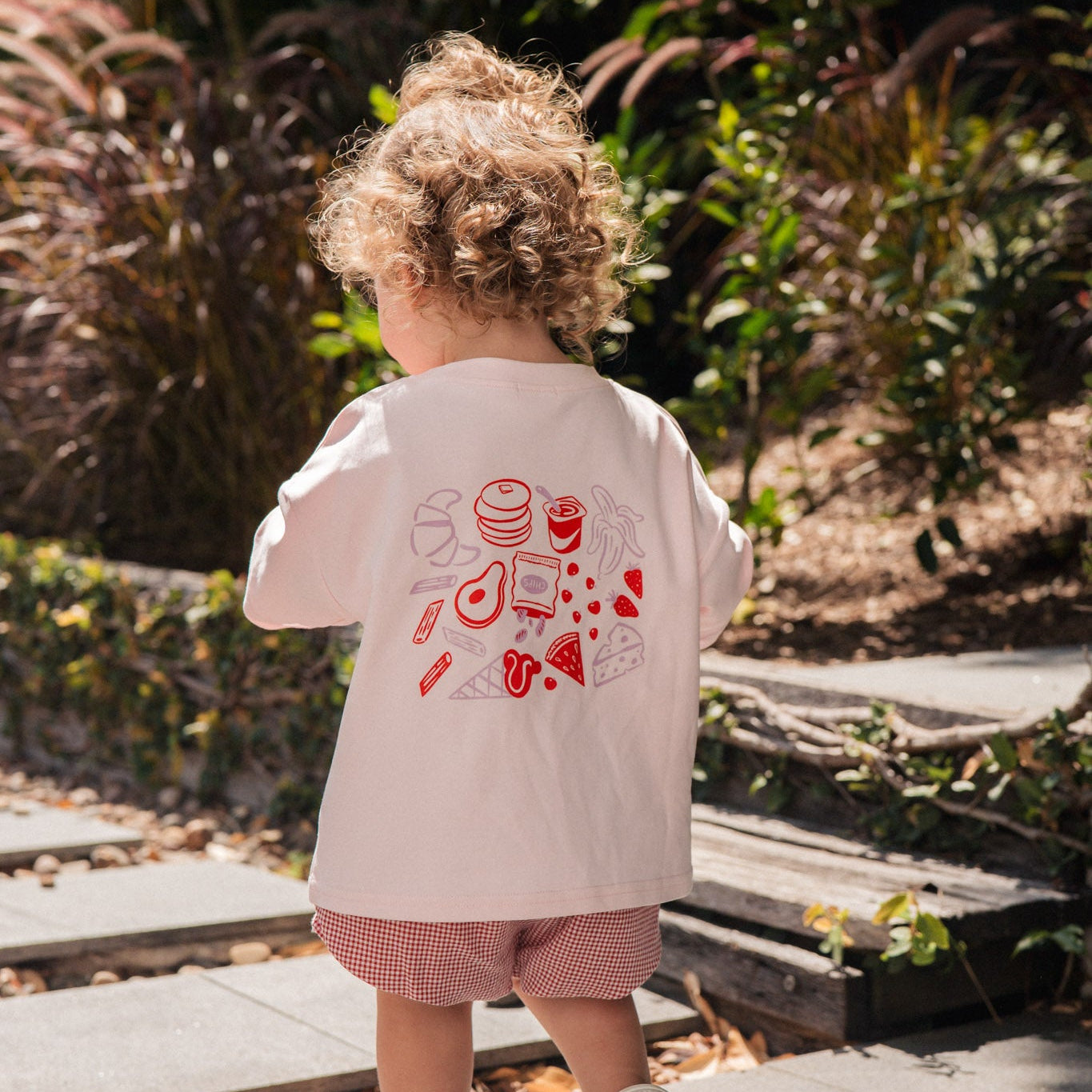 Child wearing a light pink snack club tee with red graphics and shorts, standing on a stone path with plants in the background.