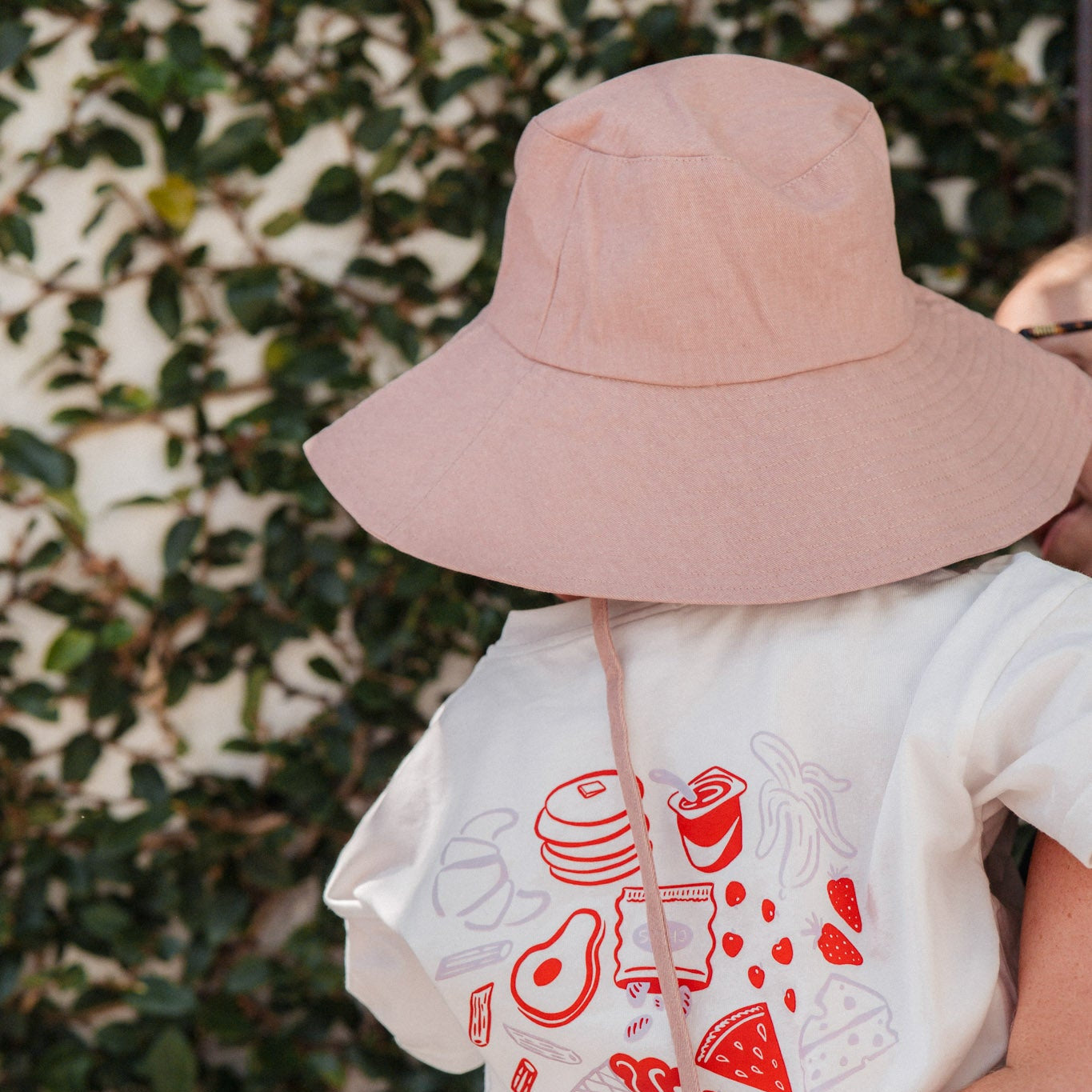 Person wearing a white t-shirt with red graphics and a pink bucket hat, standing against a leafy background.