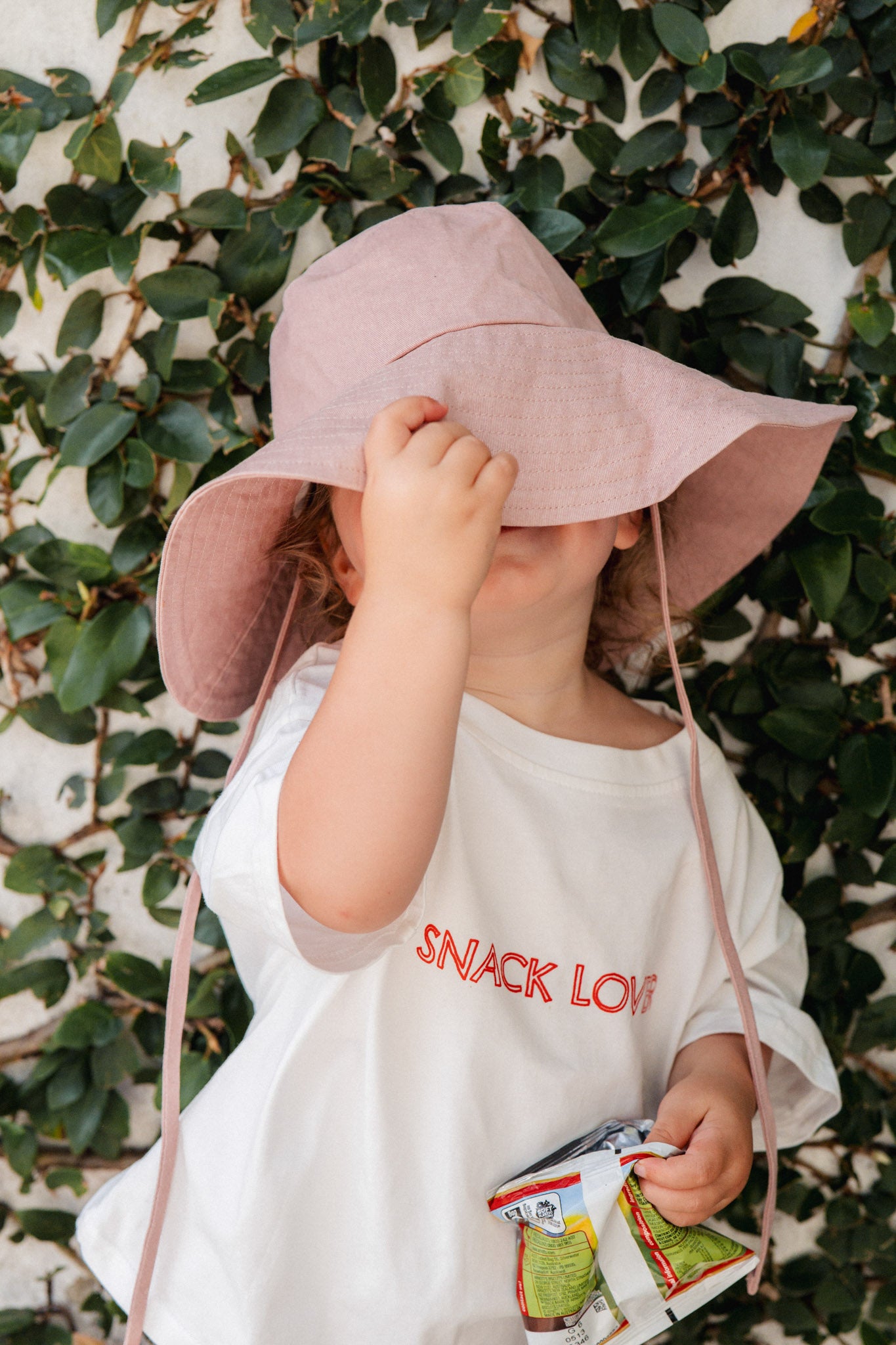 Child wearing a pink sun hat and white dress with 'SNACK LOVER' text, holding snacks against a leafy background.