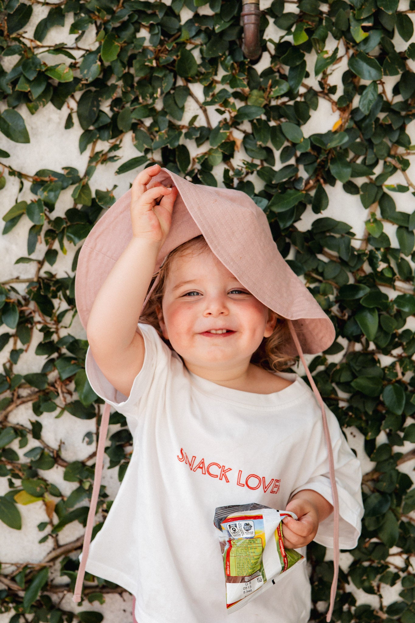 Child wearing a pink hat and white shirt with a snack design, standing against a leafy background.