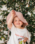 Child wearing a pink hat and white shirt with a snack design, standing against a leafy background.