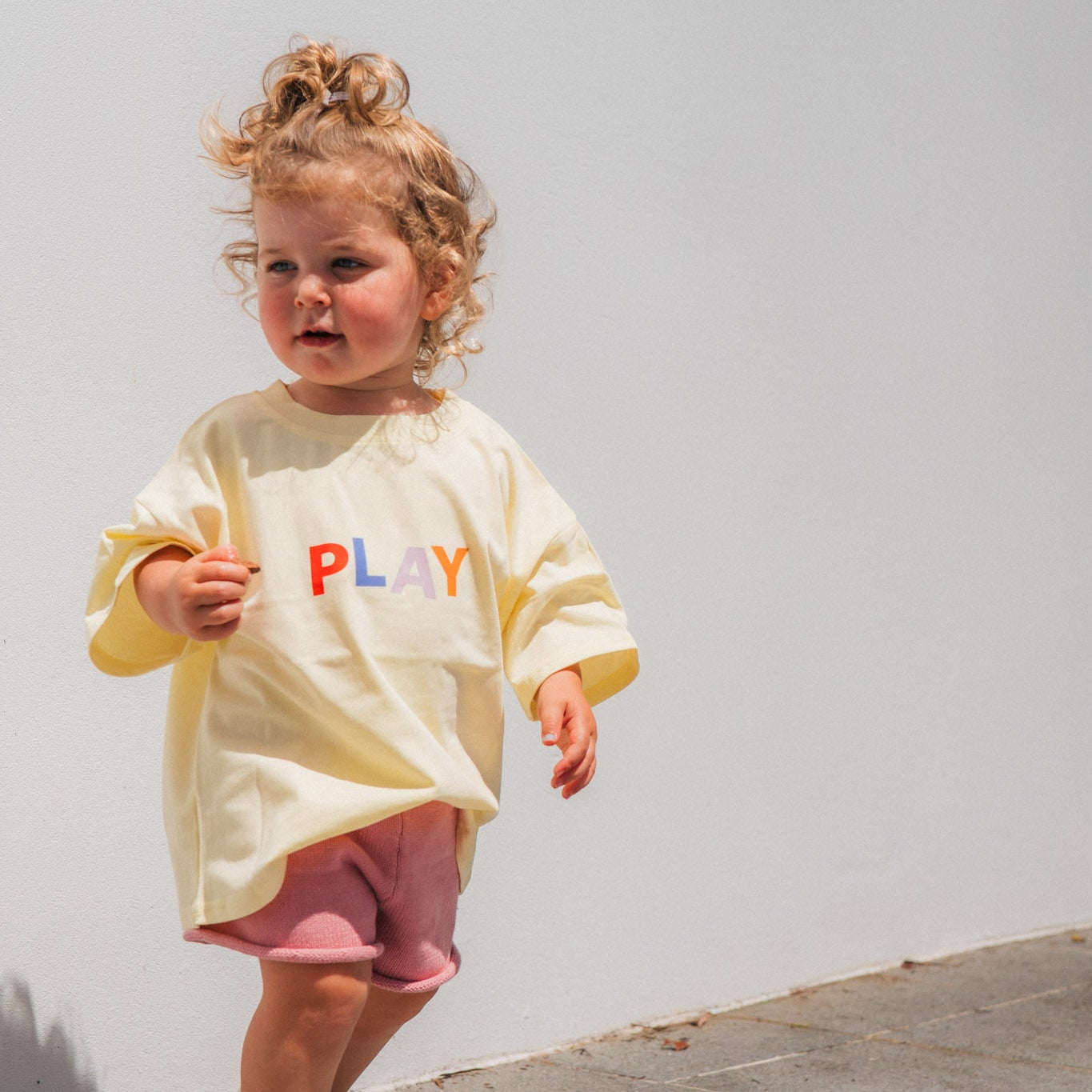 Child wearing a yellow shirt with 'PLAY' letters against a white wall.