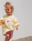 Child wearing a yellow shirt with 'PLAY' letters against a white wall.