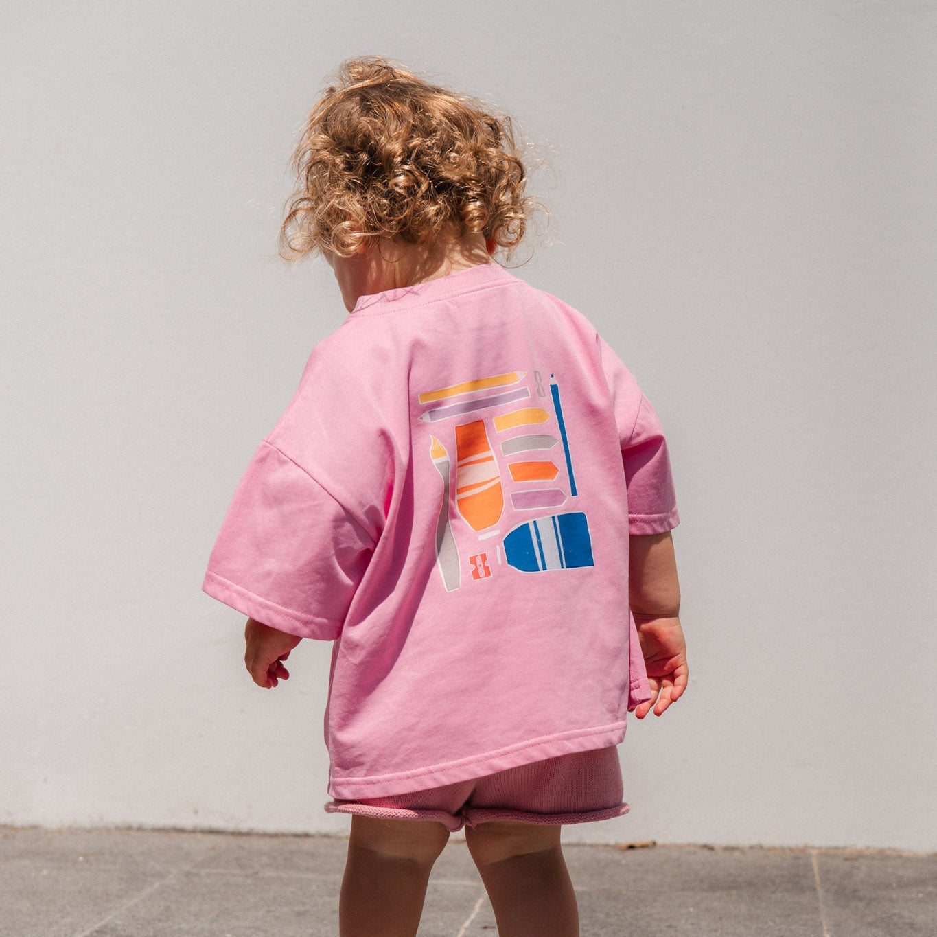 Child wearing a make more art tee with colorful designs on a gray tiled floor.