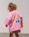Child wearing a make more art tee with colorful designs on a gray tiled floor.