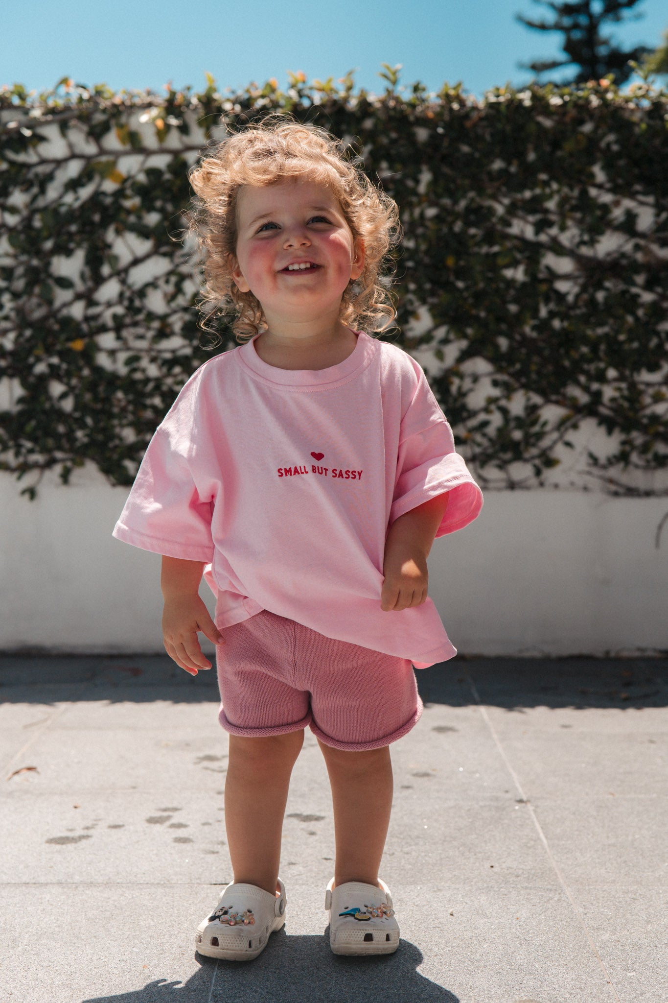 Child wearing small but sassy tee outdoors against a white wall and greenery.