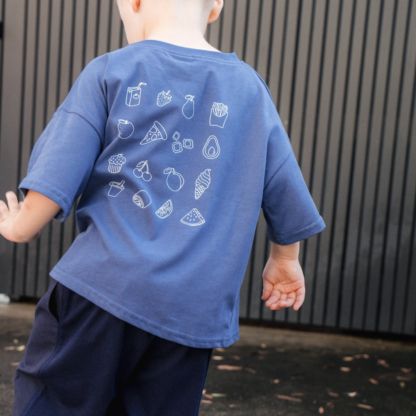Child wearing a blue t-shirt with white graphics and dark pants, standing on a pavement.