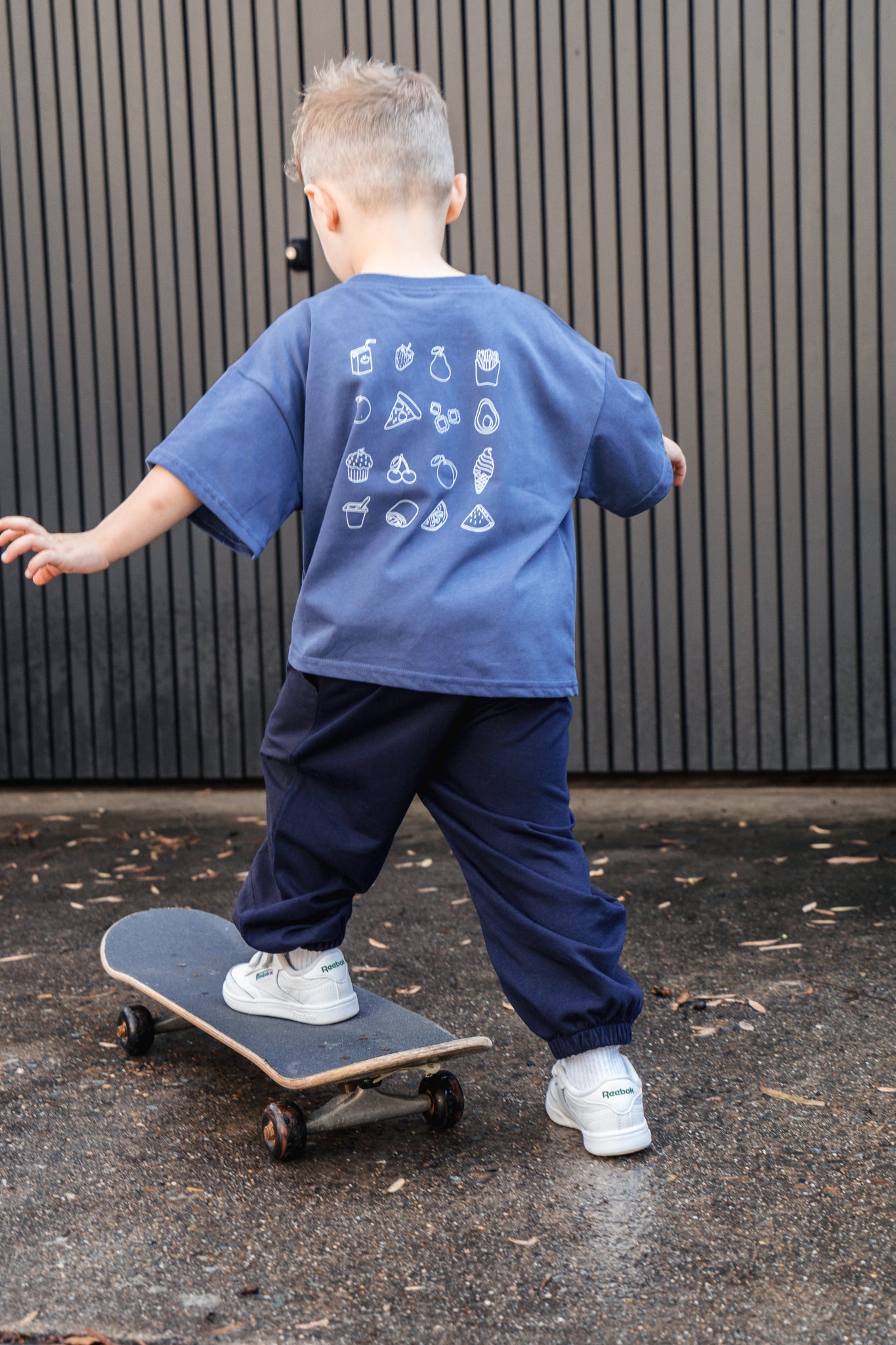 Child wearing a blue t-shirt with white graphics and dark pants, standing on a skateboard.