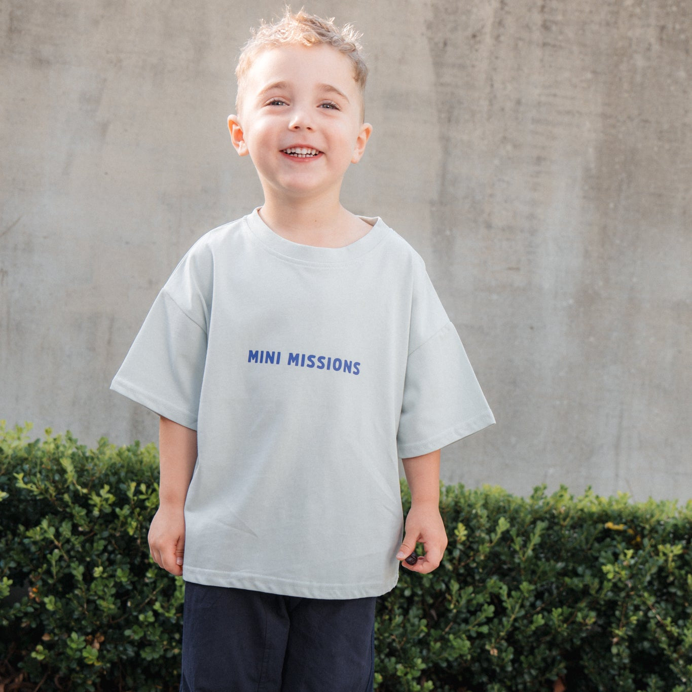 Child wearing a light gray t-shirt with 'MINI MISSIONS' text, standing outdoors against a concrete wall.