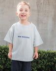 Child wearing a light gray t-shirt with 'MINI MISSIONS' text, standing outdoors against a concrete wall.
