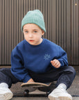 Child wearing a blue sweatshirt and green beanie sitting on a skateboard.