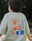 Child wearing a art t-shirt with colorful geometric design sitting on grass.