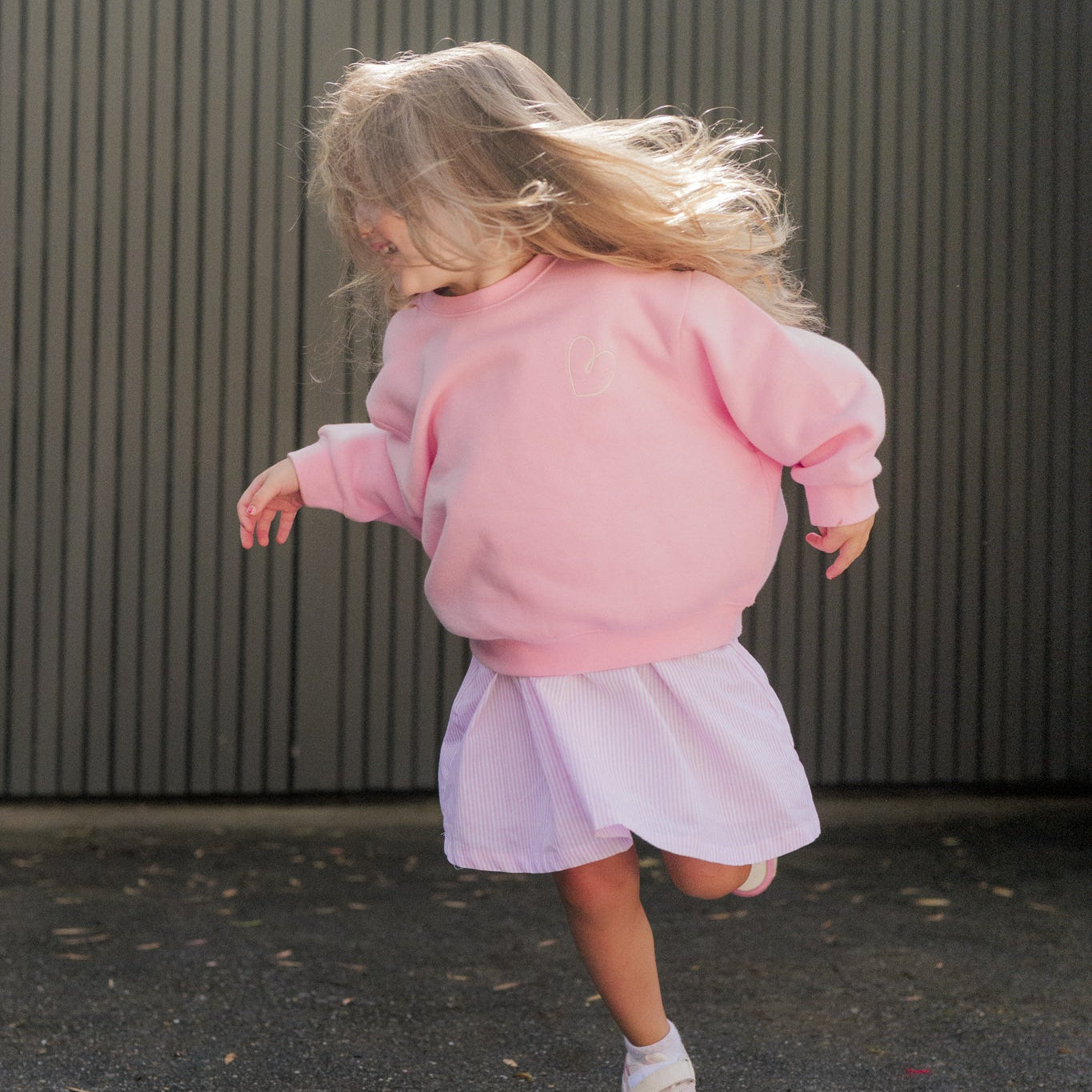 Child in a pink jumper and goldie dress running on a pavement.