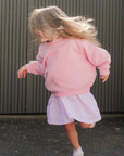 Child in a pink jumper and goldie dress running on a pavement.