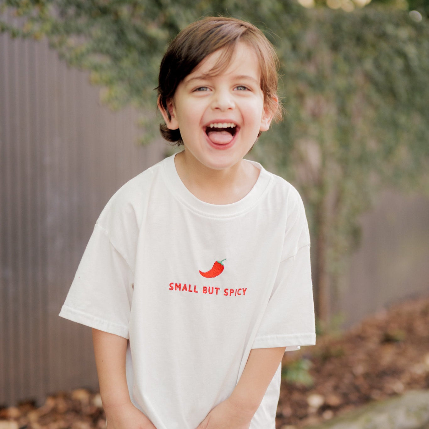 Child wearing a white t-shirt with small but spicy graphic and text, standing outdoors.