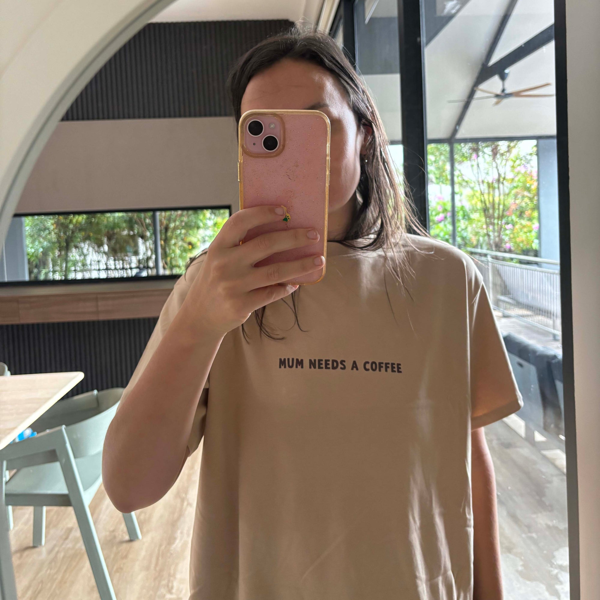 Person wearing a beige t-shirt with 'Mum needs a coffee' text, taking a mirror selfie.