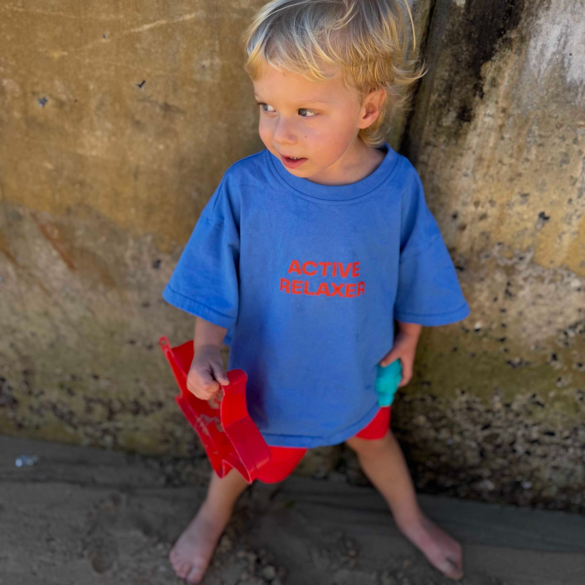 Child wearing a blue shirt with active relaxer red text and red shorts standing against a textured wall.