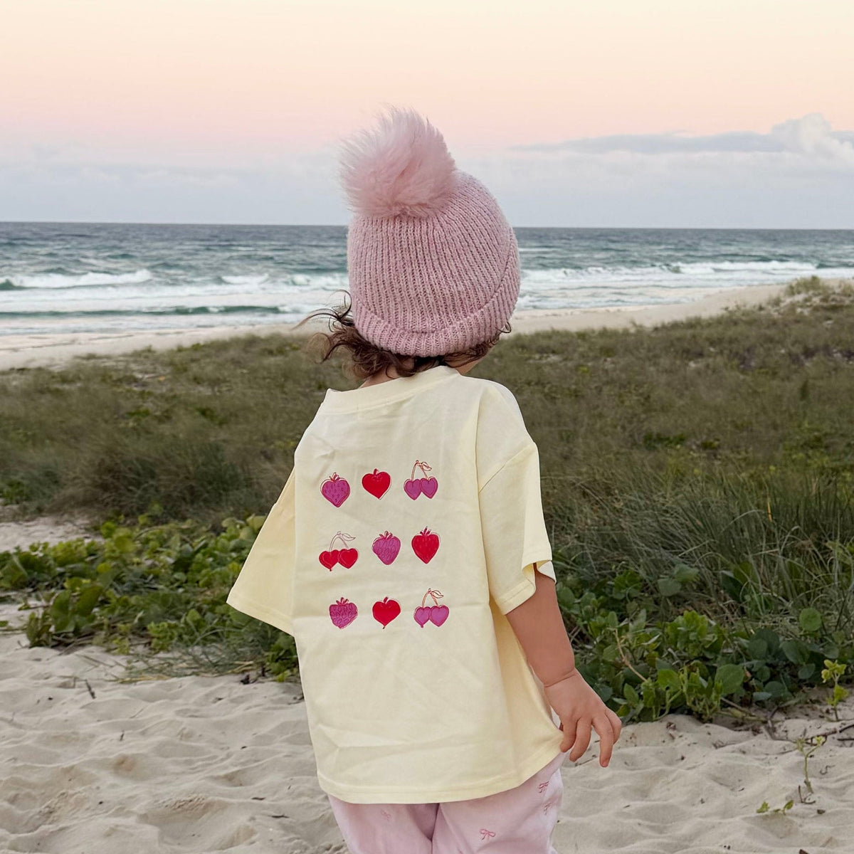 A toddler stands on a sandy beach facing the ocean at sunset, wearing a pale yellow t-shirt with a heart pattern on the back, a pink knit pom-pom beanie, and pink pants.