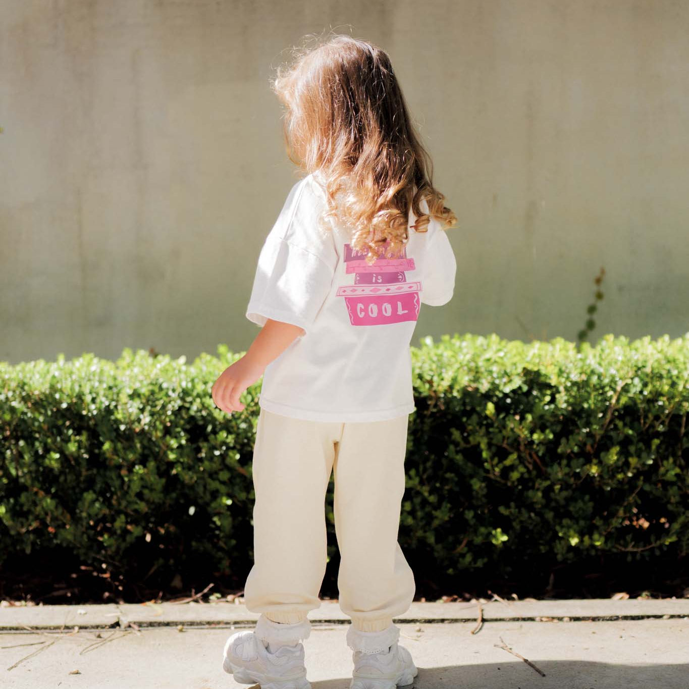 Child wearing a white shirt with text and beige pants standing on a sidewalk.
