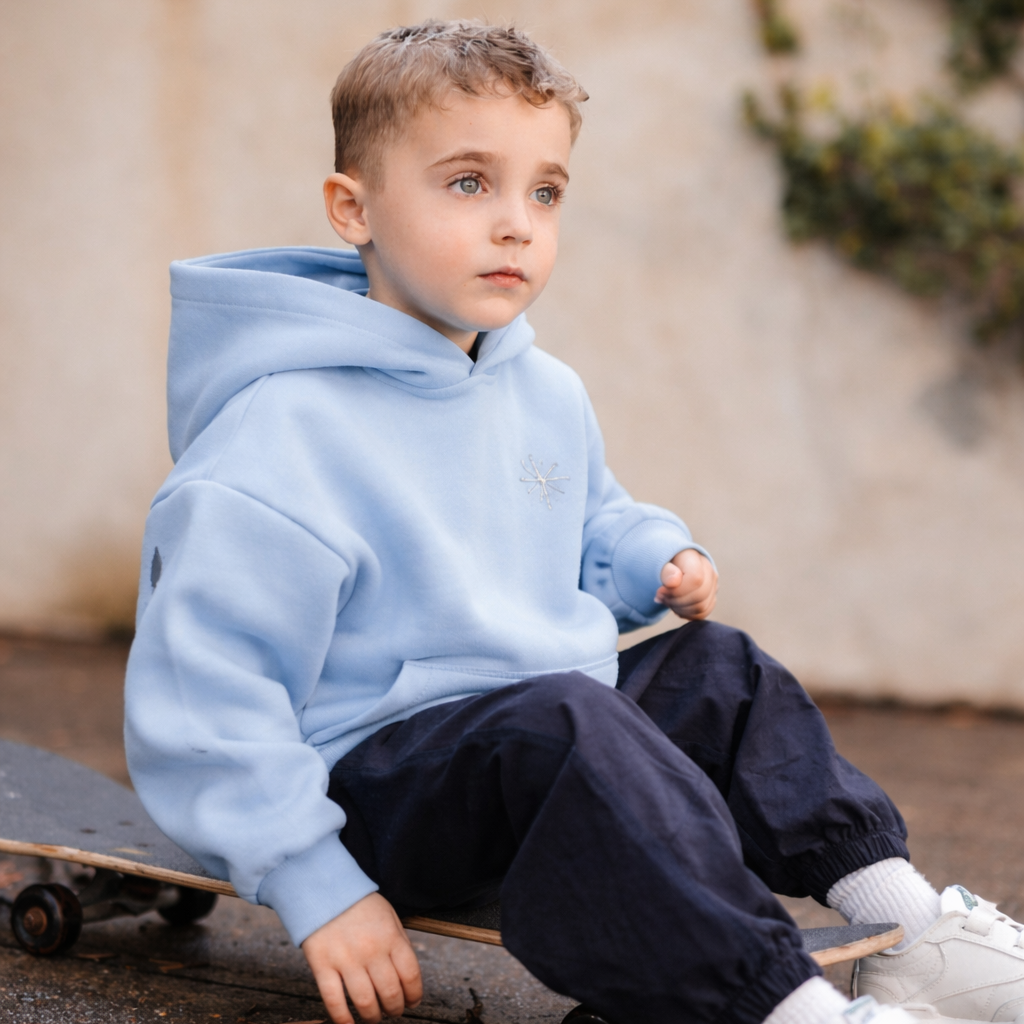 Child wearing a light blue hoodie and dark pants sitting on a skateboard.