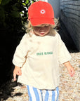 A child wearing a beige t-shirt with the phrase 'FREE RANGE' printed on the front in green, paired with striped pants and a red hat.