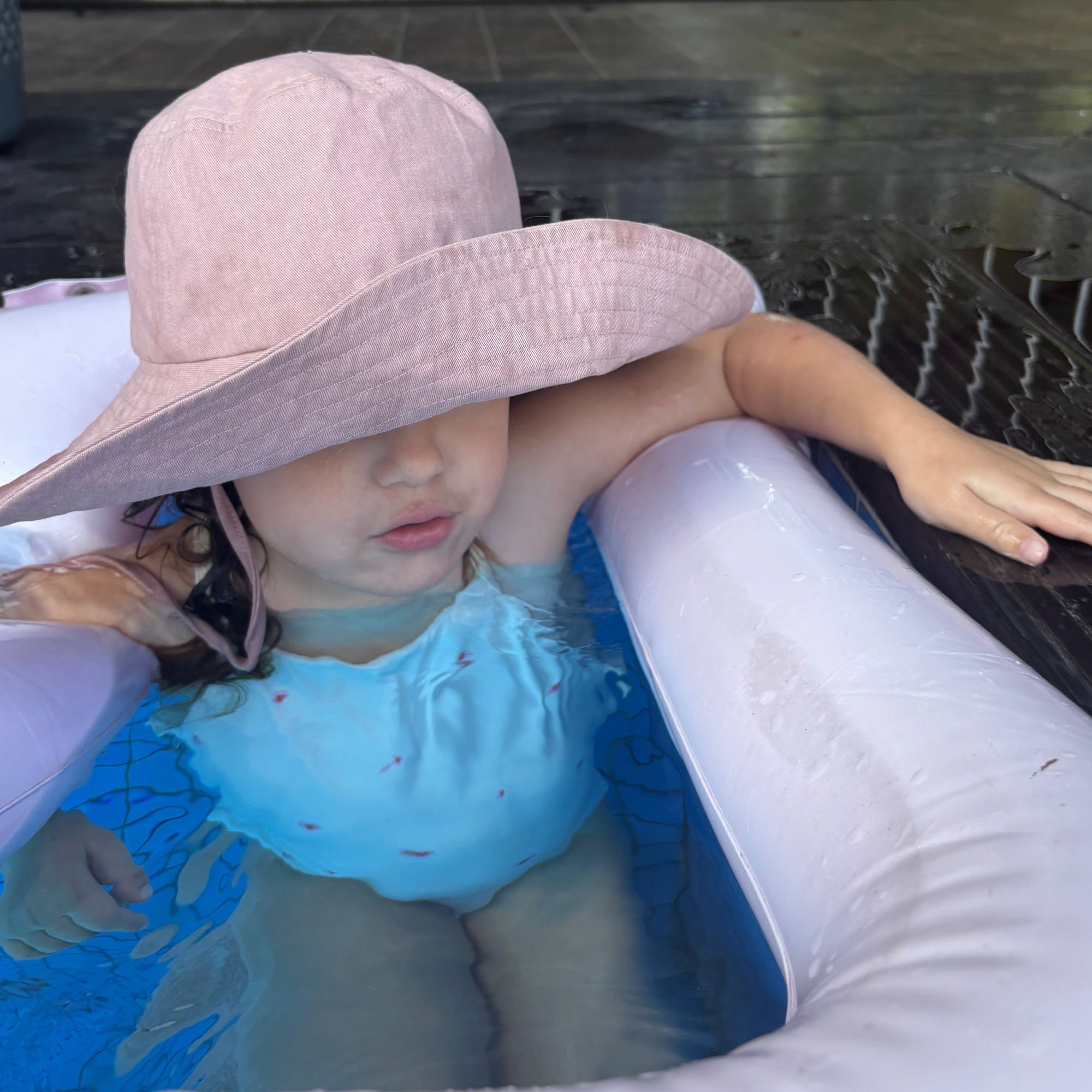 Child in a pool with a large pink hat and blue swim shirt.