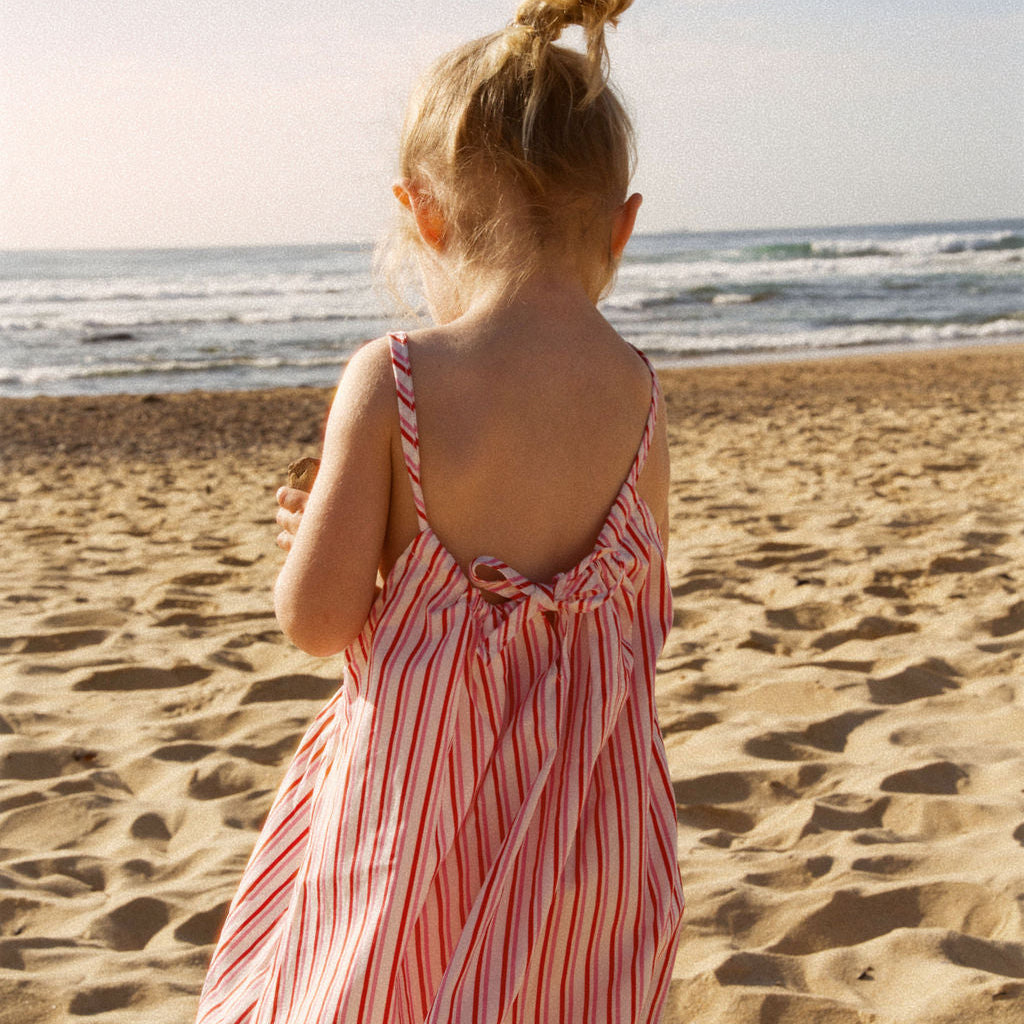 Young girl in a striped dress standing on a sandy beach looking at the ocean.