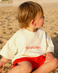Child sitting on a sandy beach wearing a white shirt with 'food critic' text and red shorts.