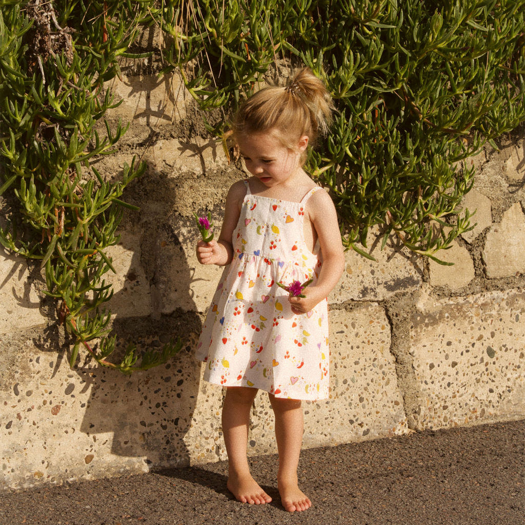 Young girl in a floral dress holding flowers against a stone wall and greenery.