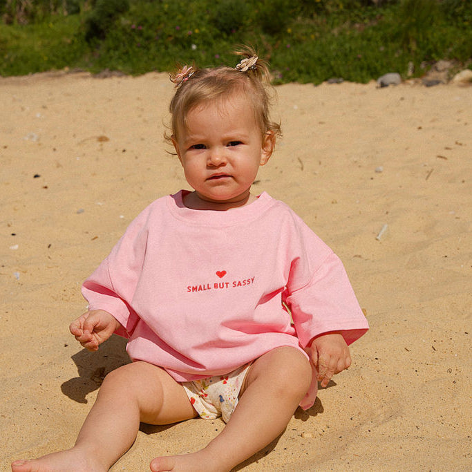 Child wearing a pink shirt with text sitting on a sandy beach.