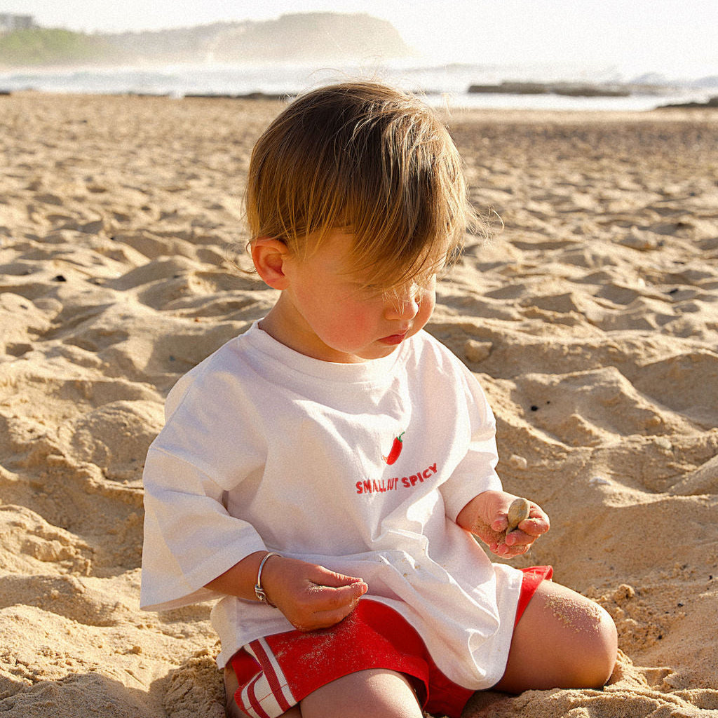 Child sitting on a sandy beach wearing a white spicy white tee and red shorts.