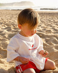 Child sitting on a sandy beach wearing a white spicy white tee and red shorts.