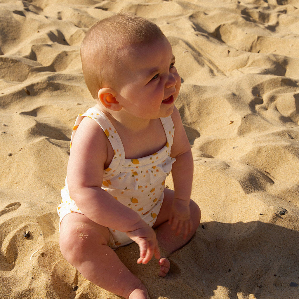 Baby sitting on sand wearing a white romper with orange flowers