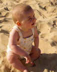 Baby sitting on sand wearing a white romper with orange flowers
