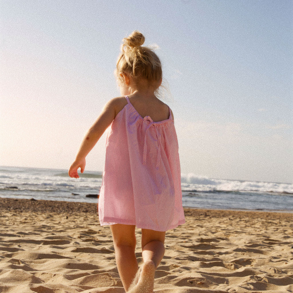 Young girl in a pink dress walking on a sandy beach with ocean waves in the background.