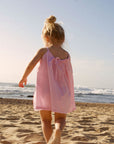 Young girl in a pink dress walking on a sandy beach with ocean waves in the background.