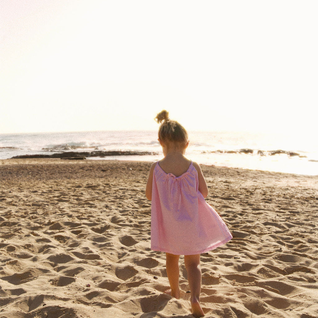 Child in a pink dress standing on a sandy beach with the ocean in the background