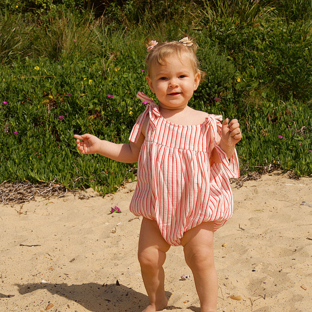 Child in a pink striped outfit standing on sandy ground with greenery in the background