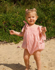 Child in a pink striped outfit standing on sandy ground with greenery in the background