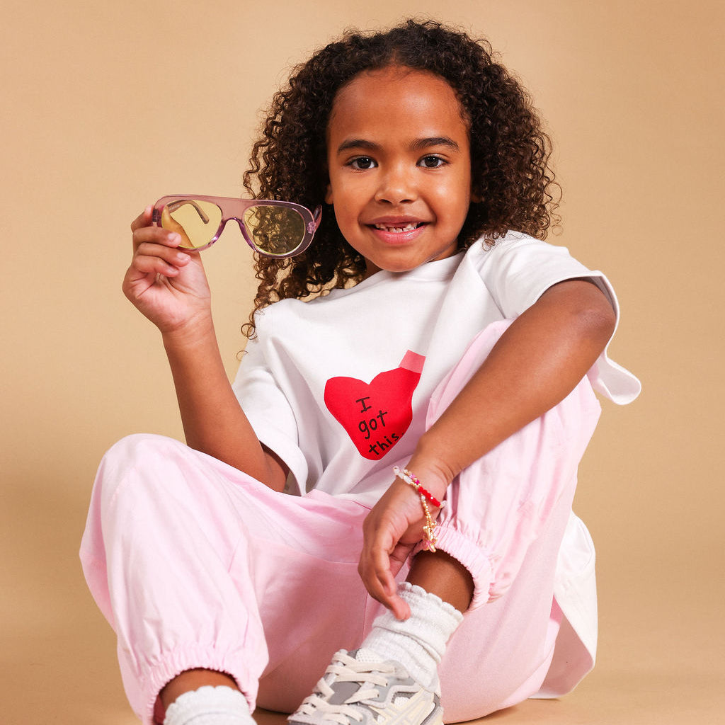 A young girl with curly hair sits on a beige studio background, wearing a white t-shirt with a red heart graphic that reads 'I GOT THIS', light pink pants, white socks, and chunky white sneakers. She is holding a pair of pink-framed sunglasses.