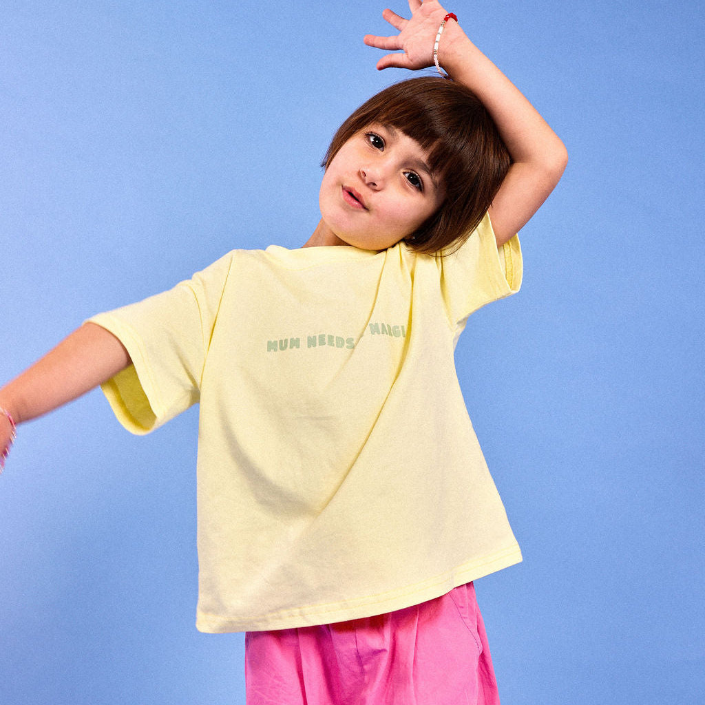 A young girl posing in a light yellow oversized t-shirt with mum needs a margi green text and pink pants against a solid blue background.