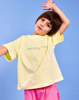 A young girl posing in a light yellow oversized t-shirt with mum needs a margi green text and pink pants against a solid blue background.