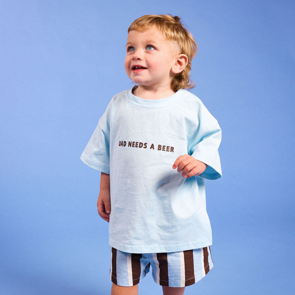 A toddler standing against a solid blue studio background, wearing a light blue oversized T-shirt with the text 'DAD NEEDS A BEER' in dark brown, striped shorts, and white sneakers.