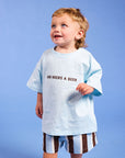 A toddler standing against a solid blue studio background, wearing a light blue oversized T-shirt with the text 'DAD NEEDS A BEER' in dark brown, striped shorts, and white sneakers.