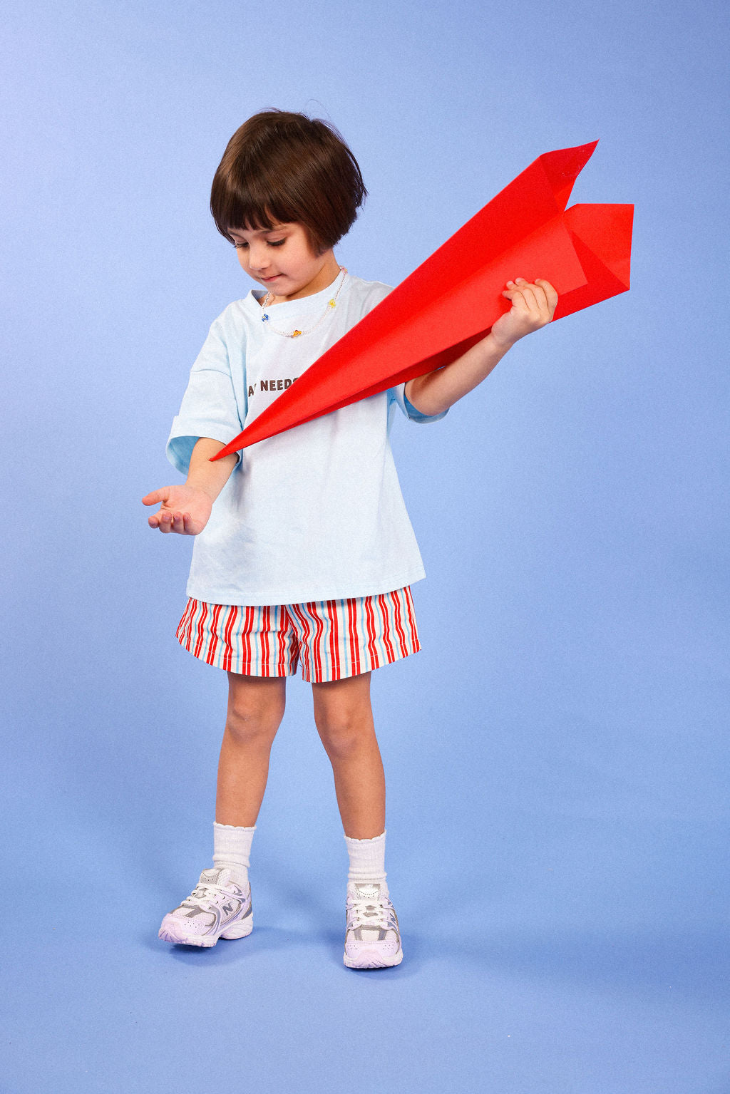 A young child stands against a light blue studio background holding a bright red paper airplane. The child is wearing a light blue T-shirt, red-and-white striped shorts, white socks, and white sneakers.