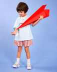 A young child stands against a light blue studio background holding a bright red paper airplane. The child is wearing a light blue T-shirt, red-and-white striped shorts, white socks, and white sneakers.