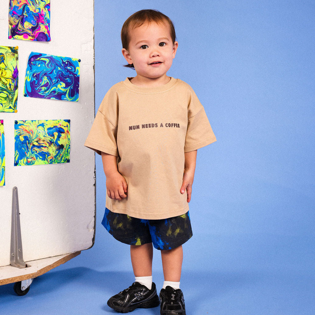 A toddler stands against a blue studio background wearing a beige oversized T-shirt with the text 'MUM NEEDS A COFFEE' and dark tie-dye shorts. A white panel with colorful abstract art is visible to the left.