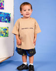A toddler stands against a blue studio background wearing a beige oversized T-shirt with the text 'MUM NEEDS A COFFEE' and dark tie-dye shorts. A white panel with colorful abstract art is visible to the left.